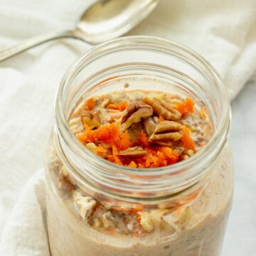 A mason jar filled with carrot cake overnight oats. Shredded carrots and pecans garnish the top. A white napkin and spoon are in the background.