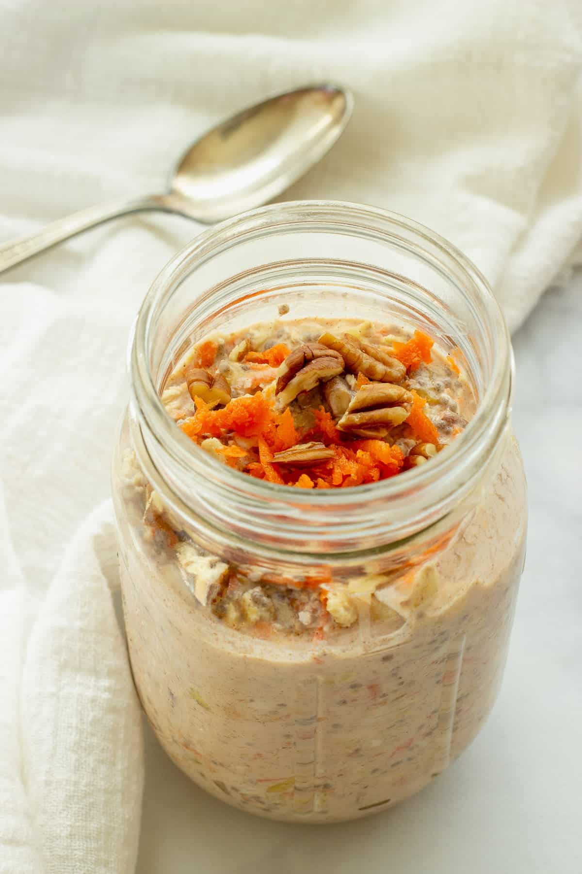 a mason jar filled with carrot cake overnight oats. Carrots and pecans garnish the top. A white napkin and spoon are in the background.