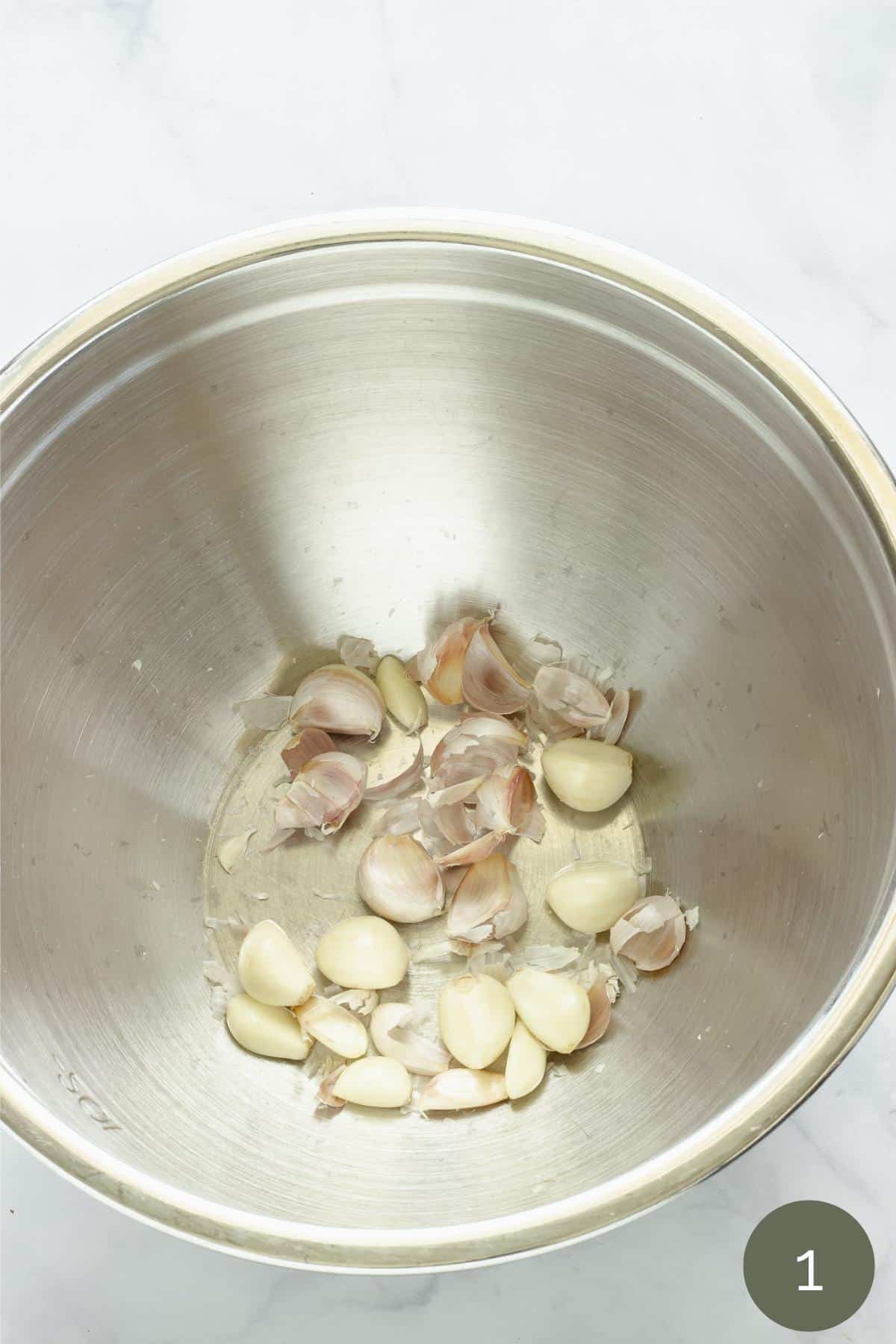 A metal mixing bowl full of garlic cloves with their skin peeled off.