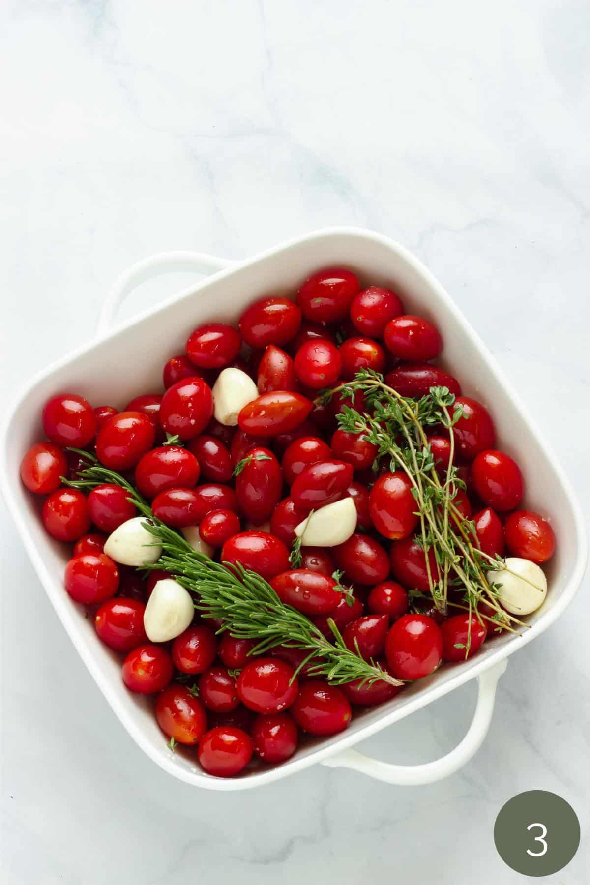 A baking dish with grape tomatoes, garlic cloves, a sprig of fresh rosemary and a few sprigs of fresh thyme.