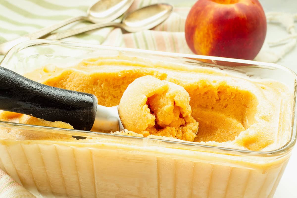 A glass loaf pan filled with peach sorbet, An ice cream scoop is resting on top with a scoop of the sorbet.