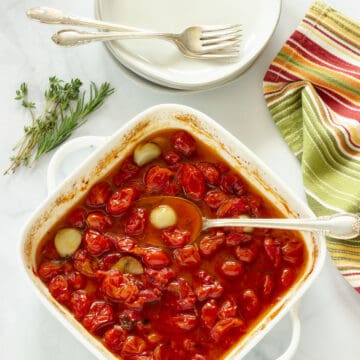 A baking dish with slow roasted grape tomatoes with garlic. A stack of white plates with forks and a striped napkin in the background.