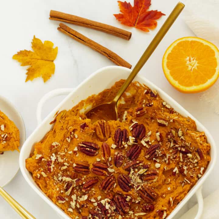 A square white casserole dish with sweet potato souffle. Serving plates, utensils, fall leaves, and cinnamon sticks are in the background.