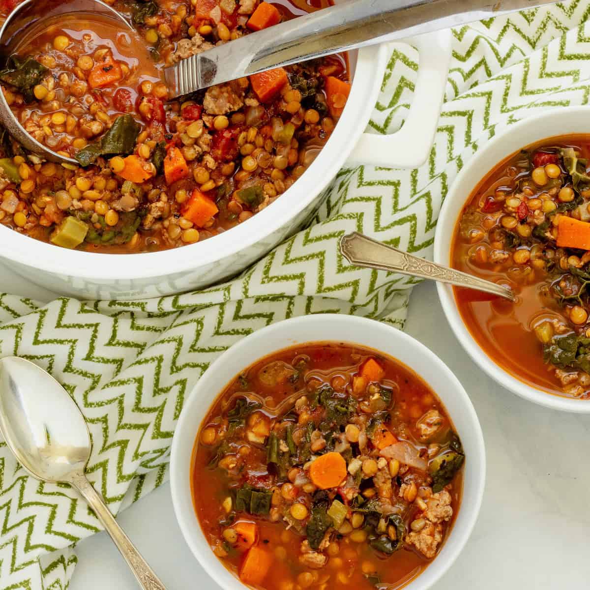 A white soup pot with ladle, filled with sausage and lentil soup, and two white soup bowls with soup. A green and white pattern napkin is in the background.
