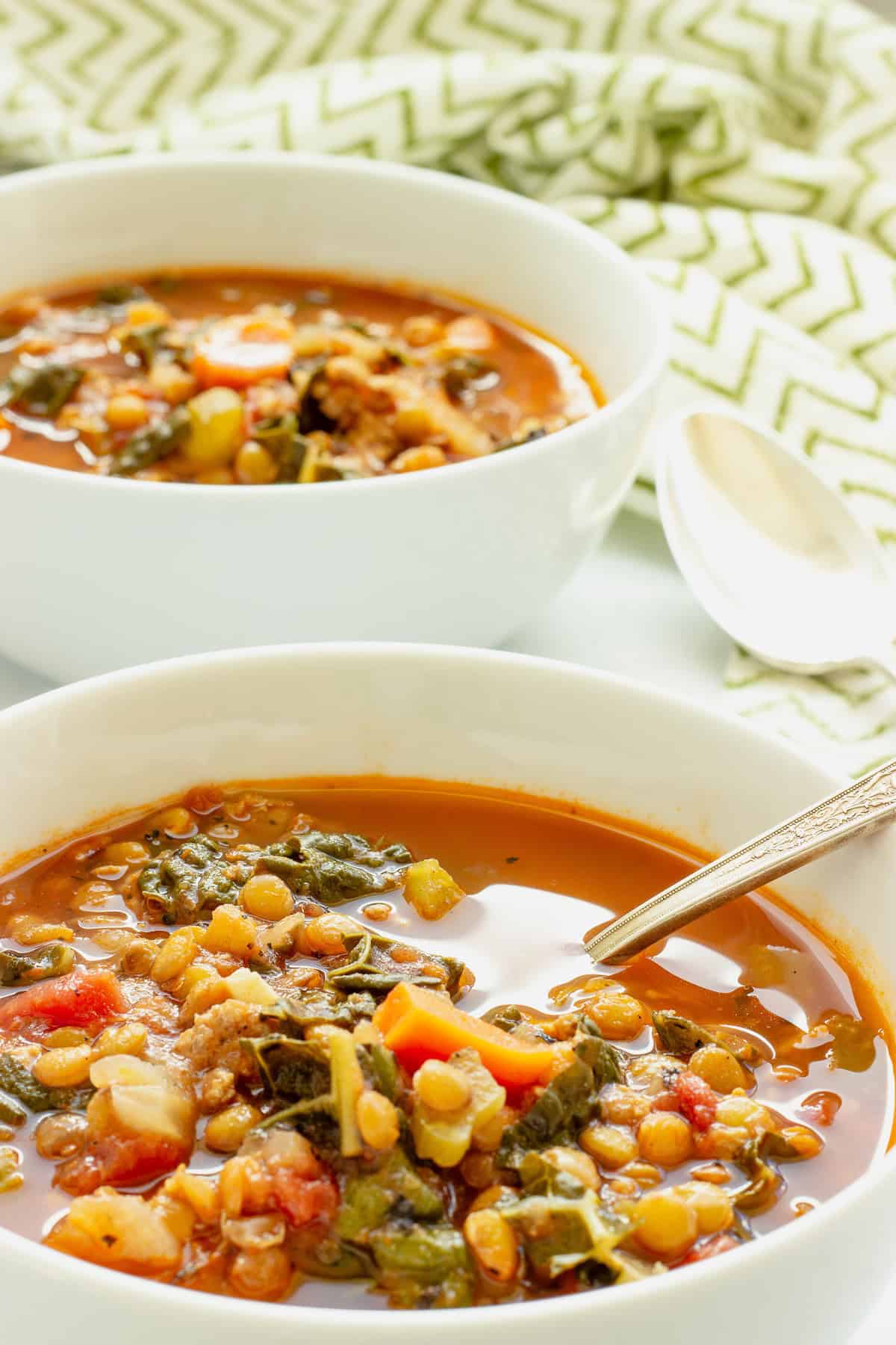 Two white bowls of sausage and lentil soup. One has a spoon in the bowl. A green and white patterned napkin is in the background.