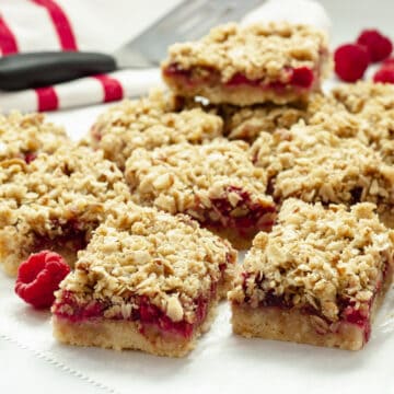 Raspberry oatmeal bars on a sheet of parchment paper with a red and white striped napkin and fresh raspberries in the background.
