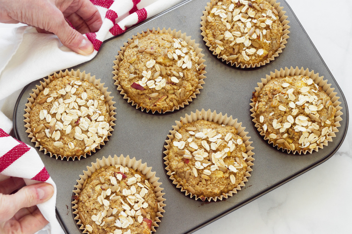 Two hands with a red and white striped napkin holding a muffin tin with six large oat bran muffins.