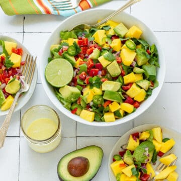 A white serving bowl with mango avocado salad. Two small serving plates with the salad, a cut avocado, and a small jar of dressing are in the background