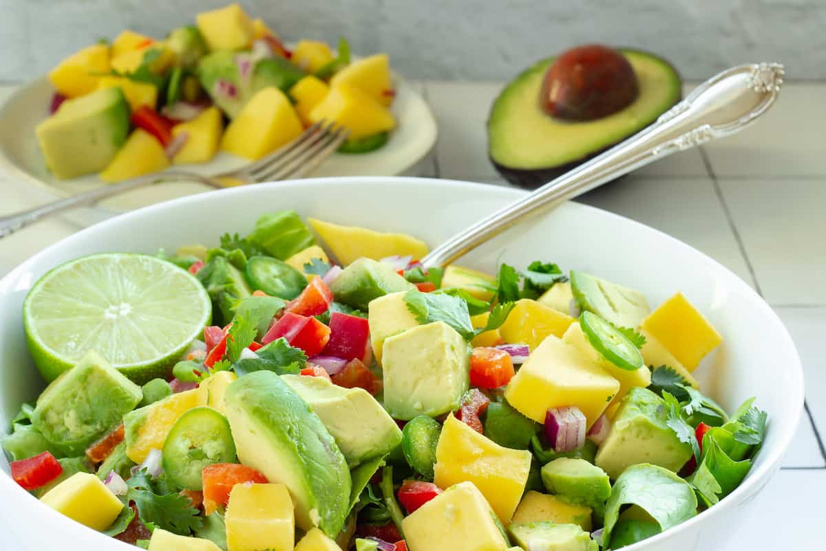 A white serving bowl with mango avocado salad and a spoon. A small serving plate and cut avocado are in the background.