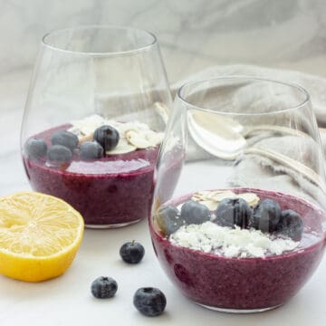 2 glasses with blueberry chia pudding garnished with fresh blueberries, coconut, and sliced almonds. A lemon is in the foreground