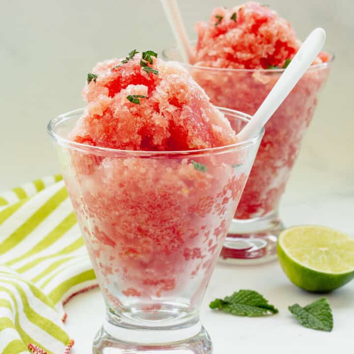Two clear glasses filled with watermelon ice. A green and white striped napkin, cut lime, and mint leaves surround the glasses.