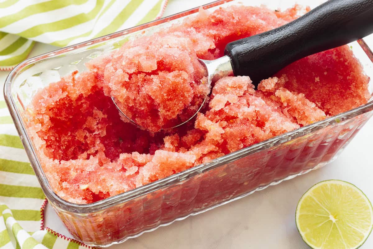 A clear loaf pan filled with watermelon ice. An ice cream scoop filled with the dessert is on top .