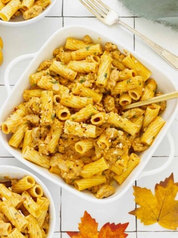 A white casserole dish with rigatoni and pumpkin pasta sauce. Small serving bowls, fall leaves, and a pumpkin are in the background.