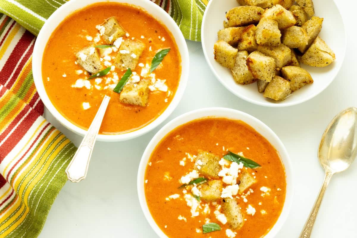 Two white bowls of roasted red pepper and tomato soup with a colorful striped napkin and a bowl of croutons.