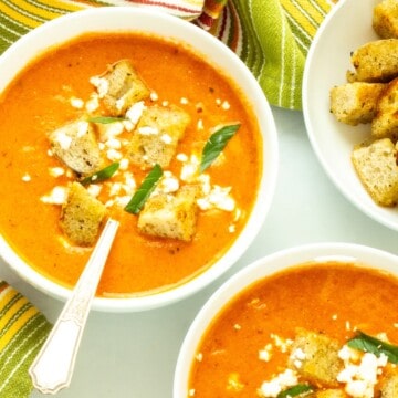 A bowl of roasted red pepper and tomato soup garnished with croutons, basil, and feta cheese.