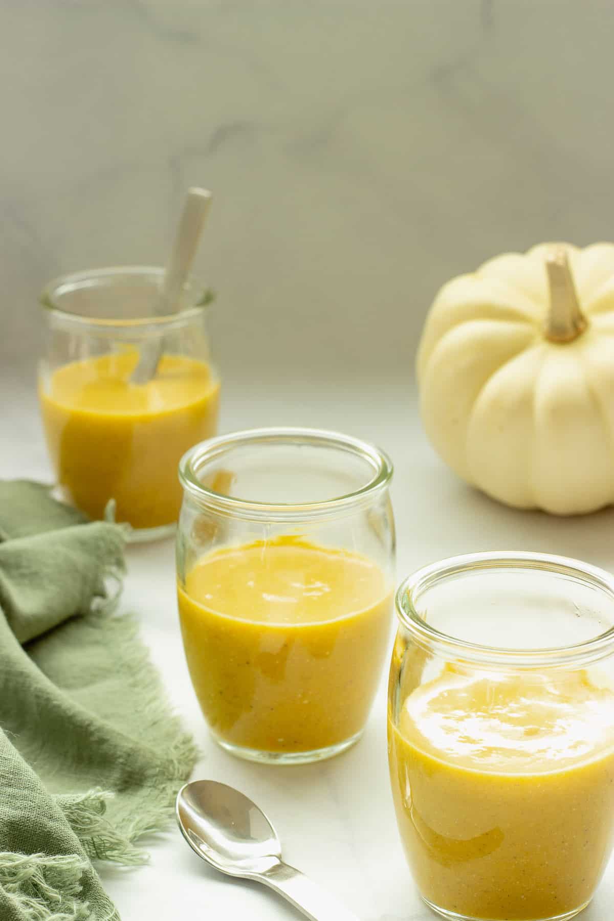 Three small shot glasses filled with spiced pumpkin bisque. A green napkin and white pumpkin are in the background.