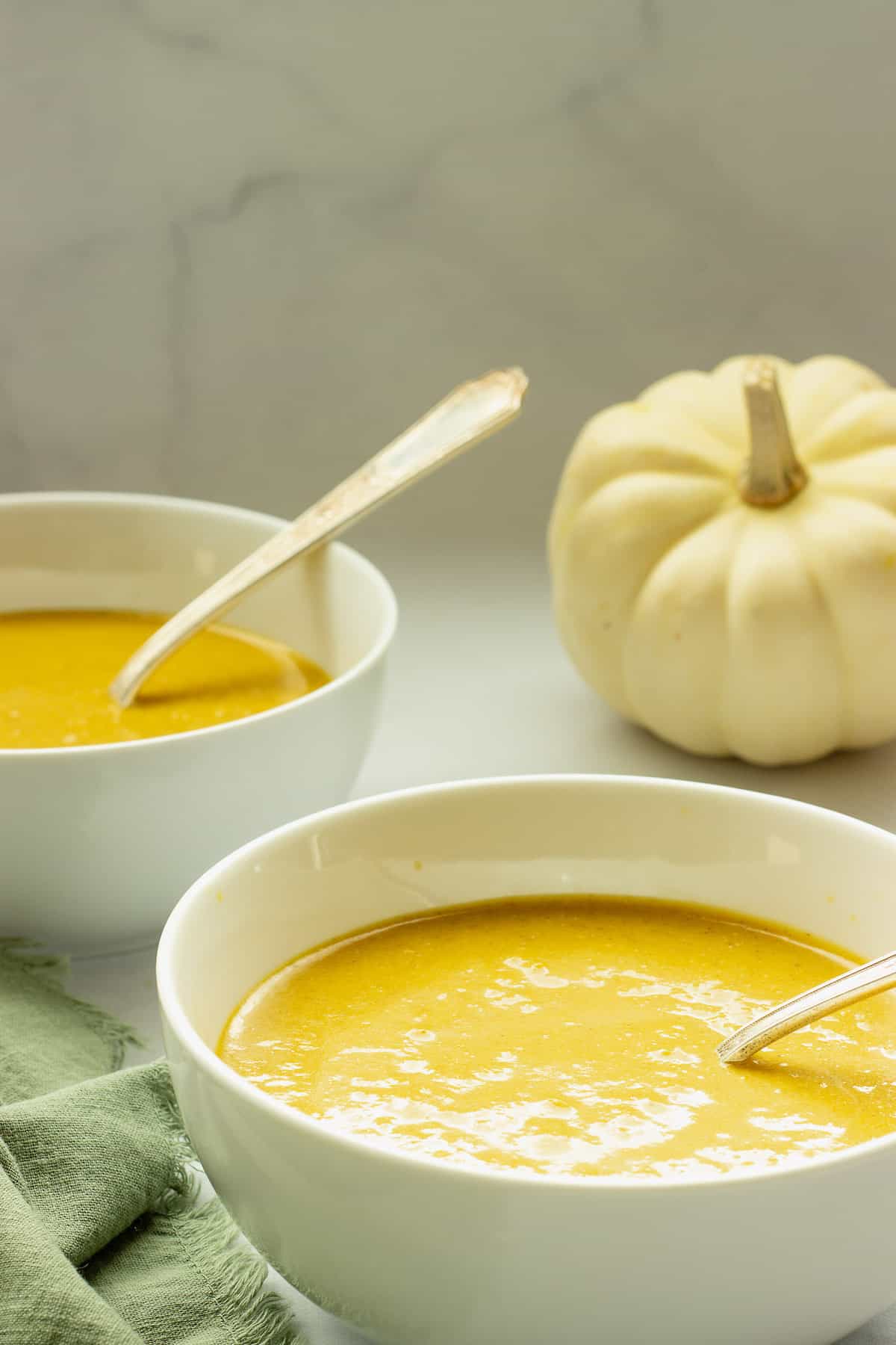 Two white bowls of spiced pumpkin bisque soup with a green napkin and white pumpkin in the background.