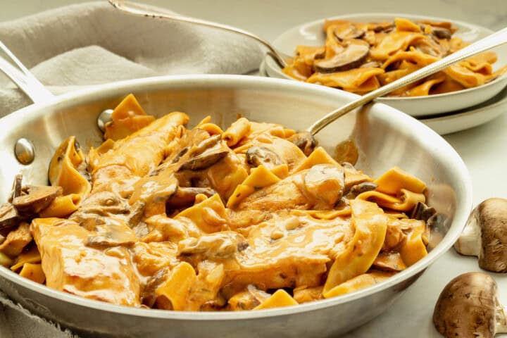A pan of creamy chicken and mushroom pasta on a white background. A plate of the dish is in the background.