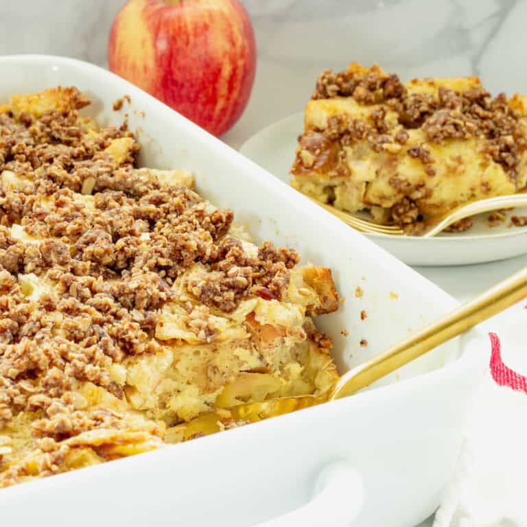 A white casserole dish with apple French toast casserole. A red apple and plate with a portion of the recipe are next to the casserole dish.
