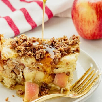 A slice of apple French toast casserole on a white plate with syrup being drizzled over the top. A red and white striped napkin and red apple are next to the plate.