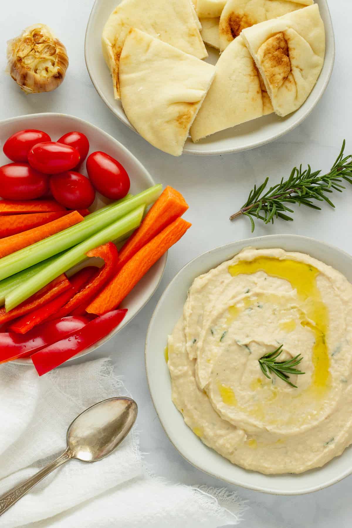 White bean dip spread on a white plate. A plate of cut vegetables and another with pita bread are in the background.