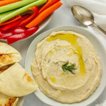 A plate spread with white bean dip. Cut vegetables and pita bread are in the background.