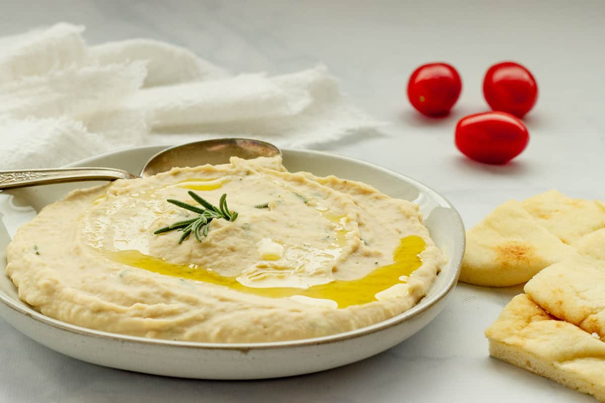A white plate with white bean dip spread onto it. Grape tomatoes and pita bread are in the background.