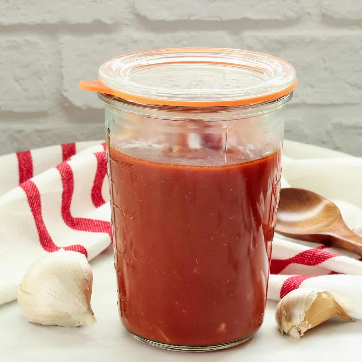 A jar of easy pasta sauce next to a red and white striped napkin, a head of garlic, and a wooden spoon.