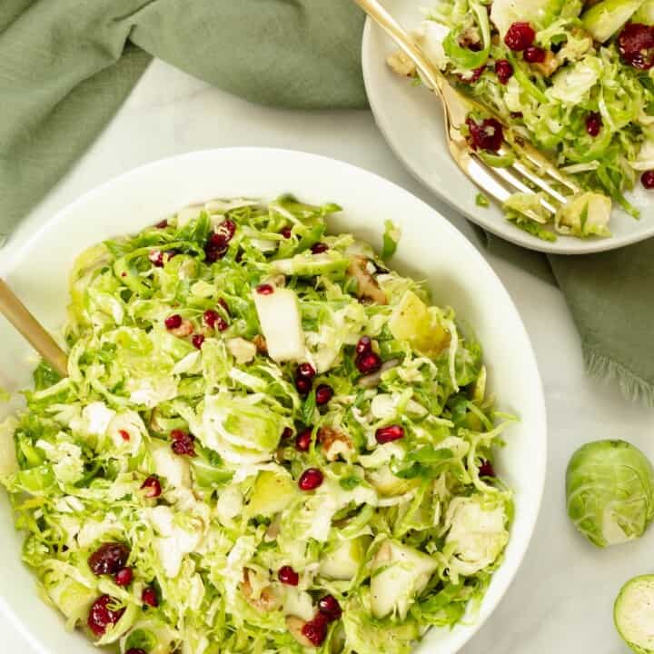 A white bowl and small plate with shaved Brussels sprouts salad. A green napkin and some raw Brussels sprouts alongside the bowl and plate.