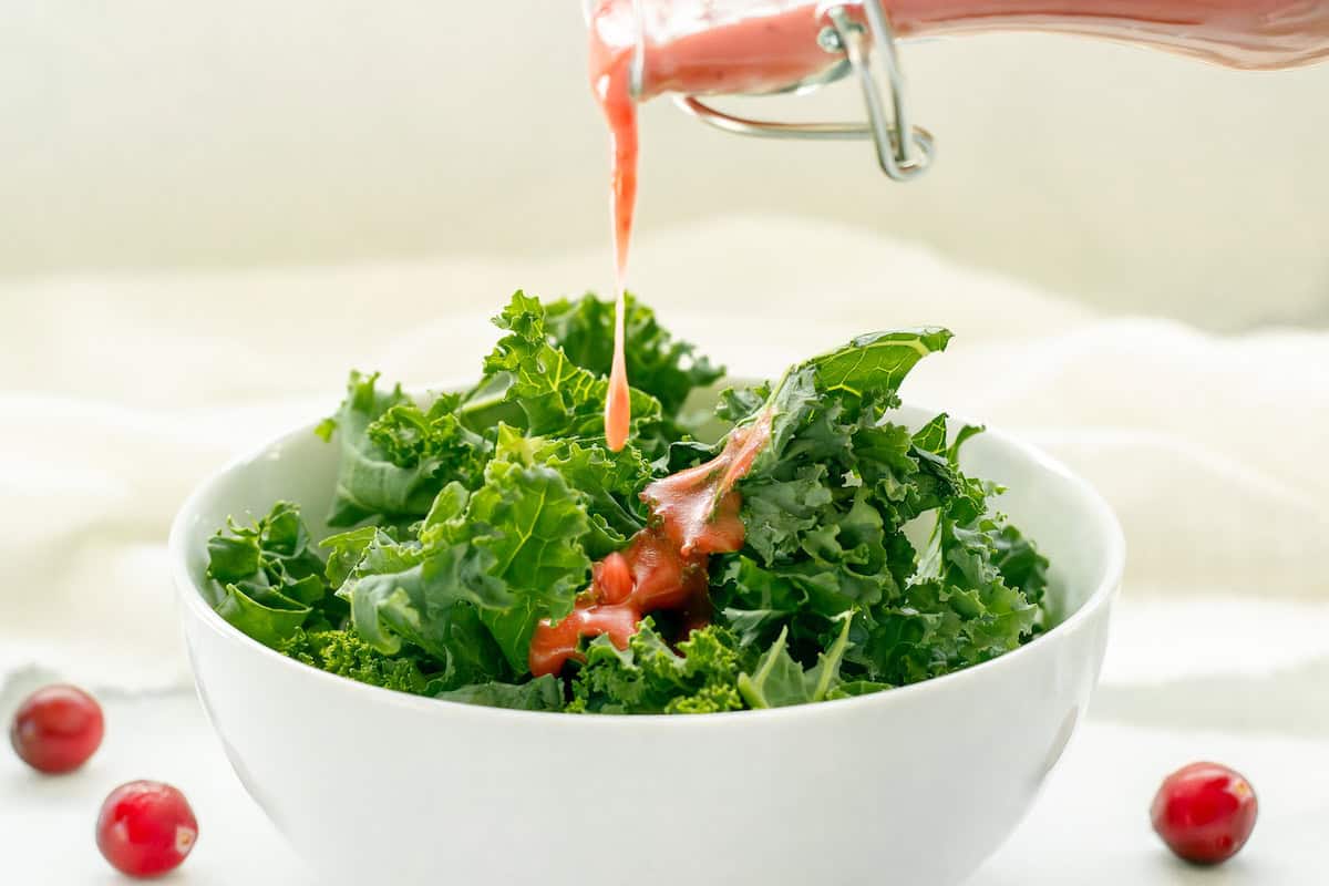 A white bowl of chopped greens. Cranberry vinaigrette is being poured over the salad.