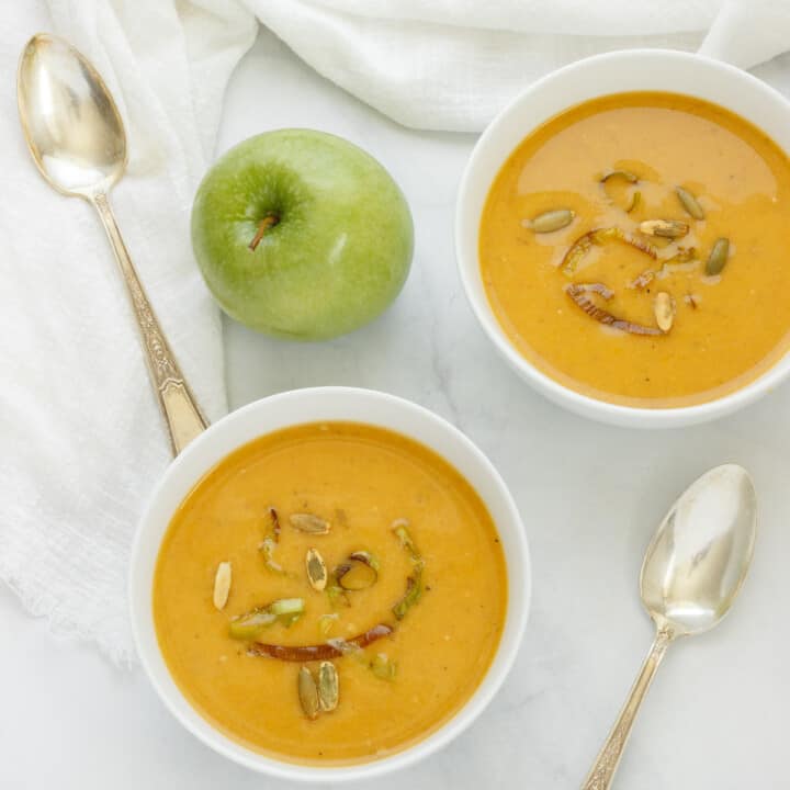 Two white bowls of butternut squash soup with apple. A green apple, spoons, and white napkin are in the background.