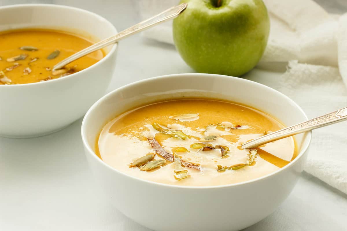 2 white bowls of butternut squash soup with apple. A green apple is in the background.