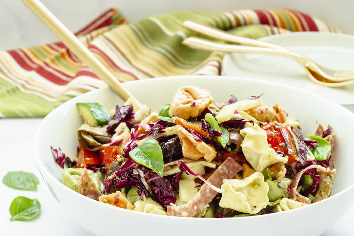 A white serving bowl filled with pesto tortellini salad. A serving spoon is in the bowl and a plate, forks, and striped napkin are in the background