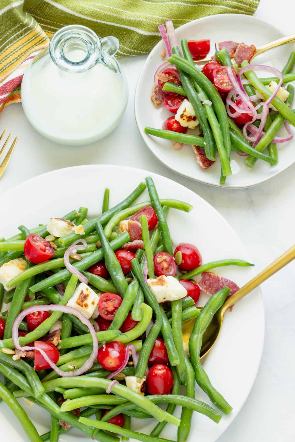 A large white plate with cold green bean salad on a white background. A bottle of salad dressing and a smaller serving plate are in the background.  
