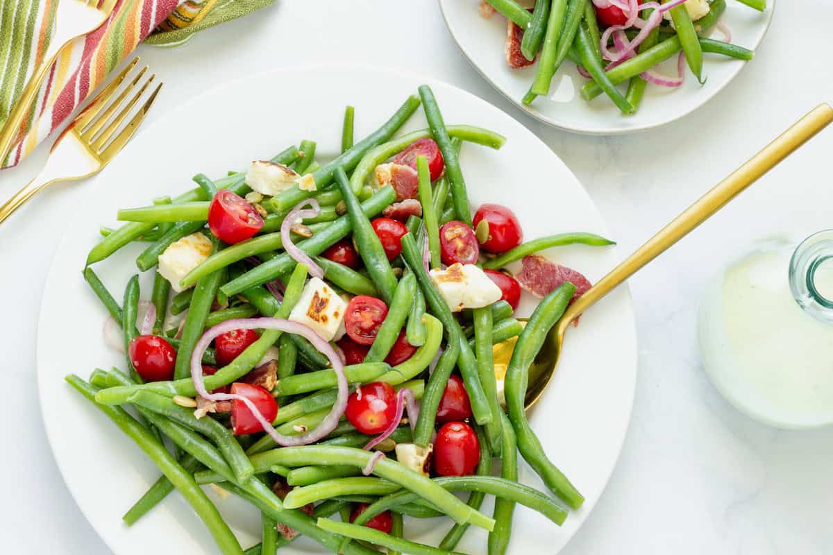 A white serving plate with cold green bean salad on a white background. A striped napkin and a small serving plate with green bean salad are in the background.