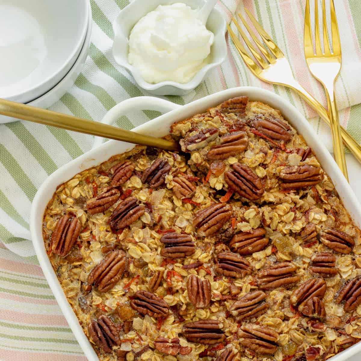 A white casserole pan with carrot cake baked oatmeal. White plates, gold forks, and a serving spoon are in the background.