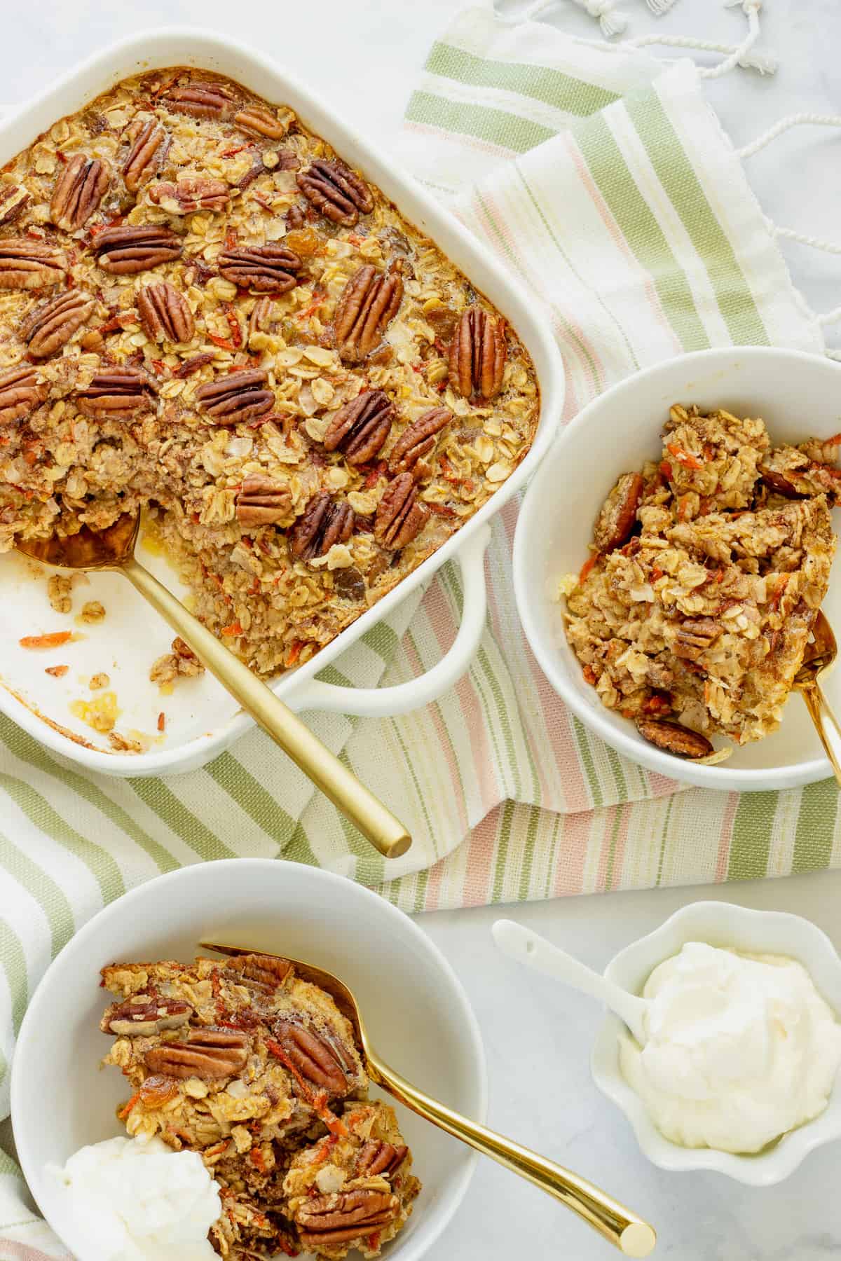 A white casserole dish with carrot cake baked oatmeal. Two bowls of the oatmeal and a small bowl with Greek yogurt cream cheese topping are in the background.