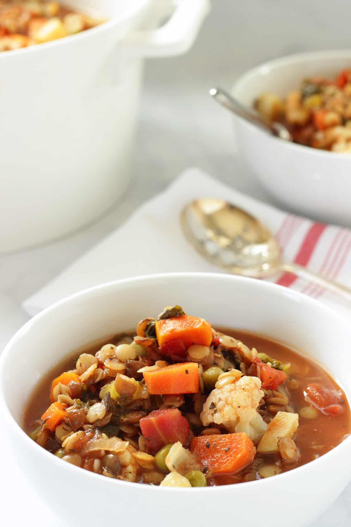 A white bowl of Heart healthy Mediterranean vegetable lentil soup. A red and white striped napkin and spoon are in the background.