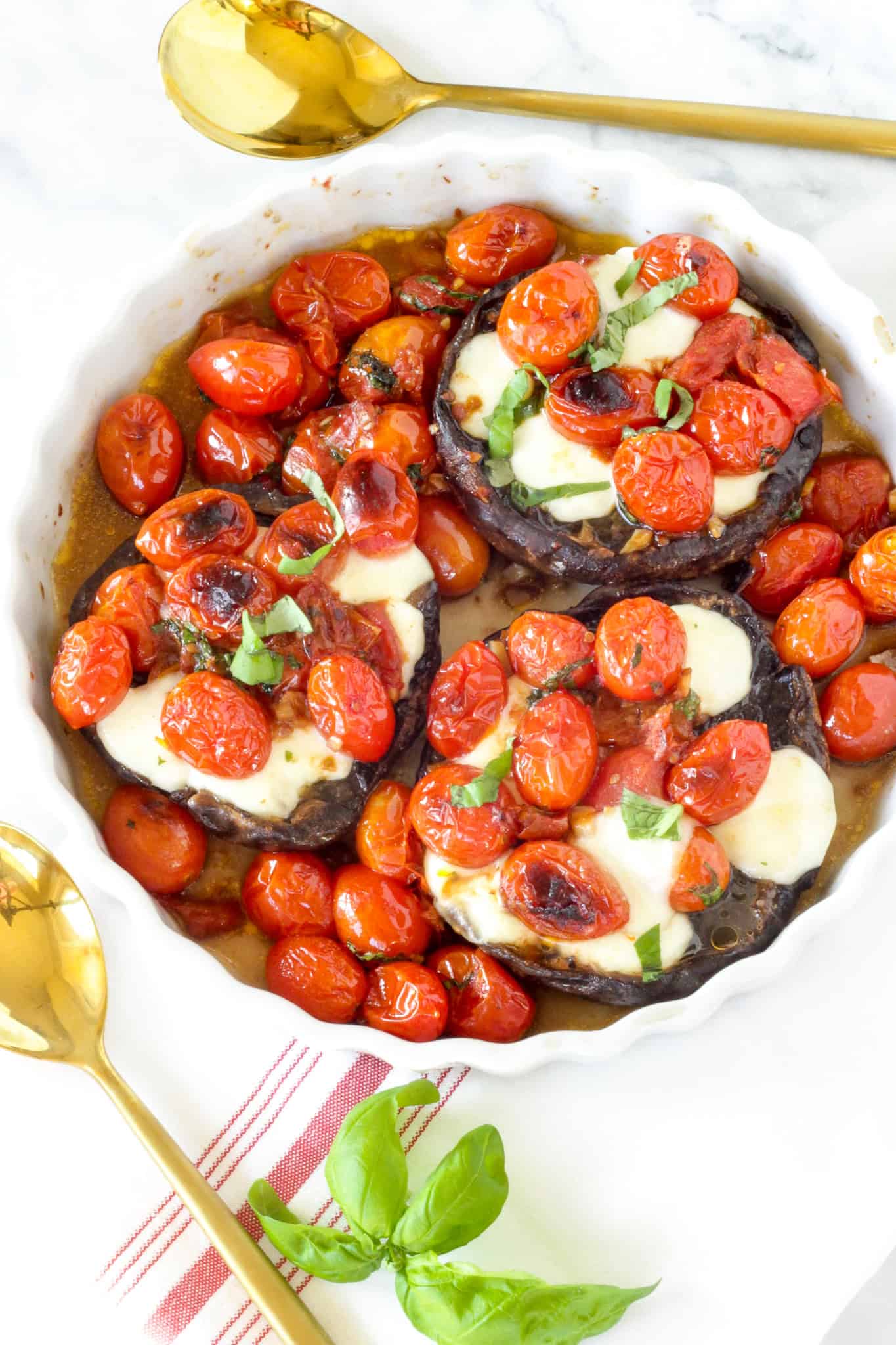 A casserole dish with Caprese Stuffed Mushrooms - three portabello mushroom caps stuffed with tomatoes and mozzarella cheese. on a white surface with red and white striped napkin.