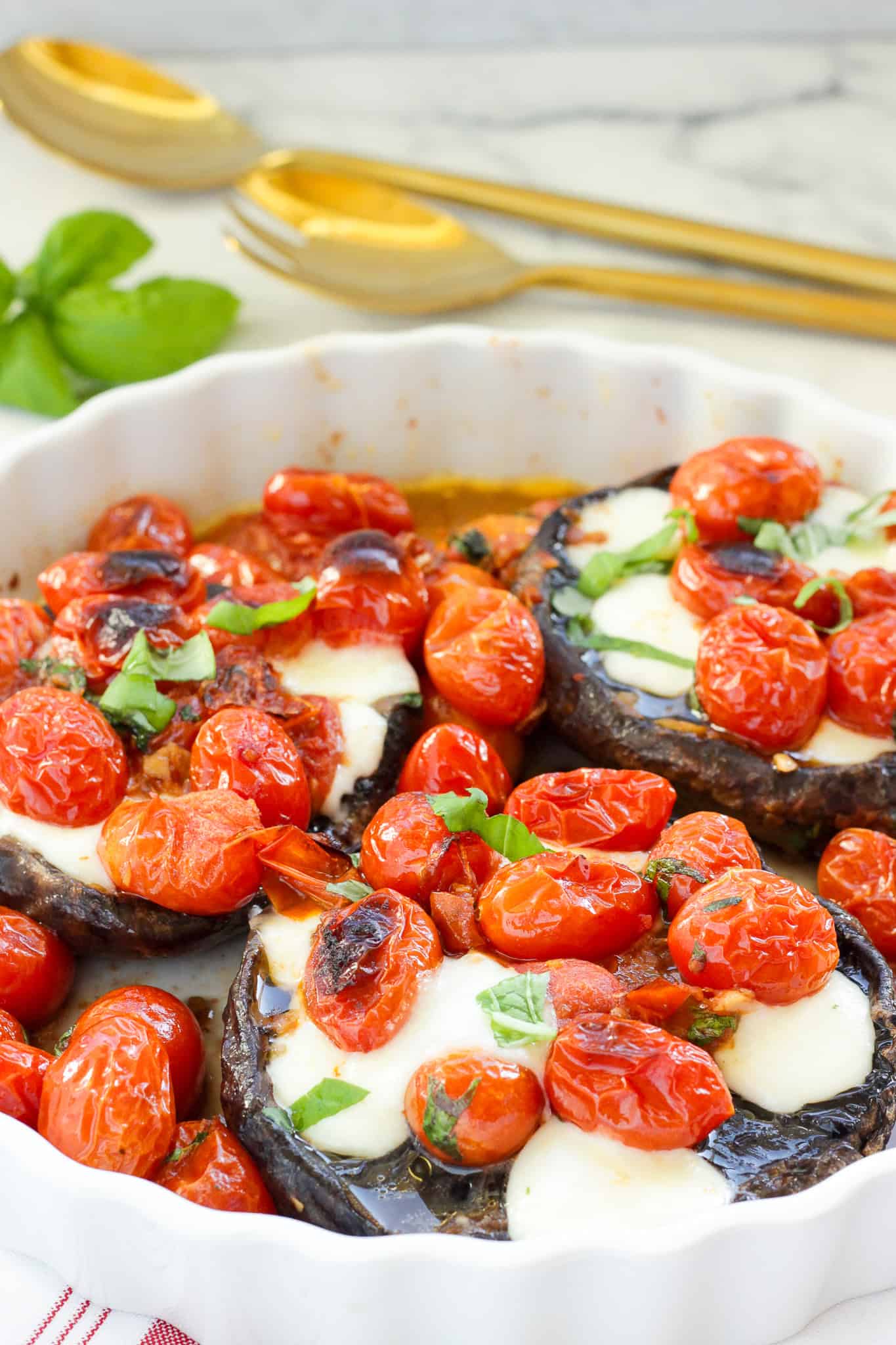 A white casserole dish with roasted Caprese Stuffed Portobello Mushrooms. Gold serving spoons and fresh basil are next to the dish.