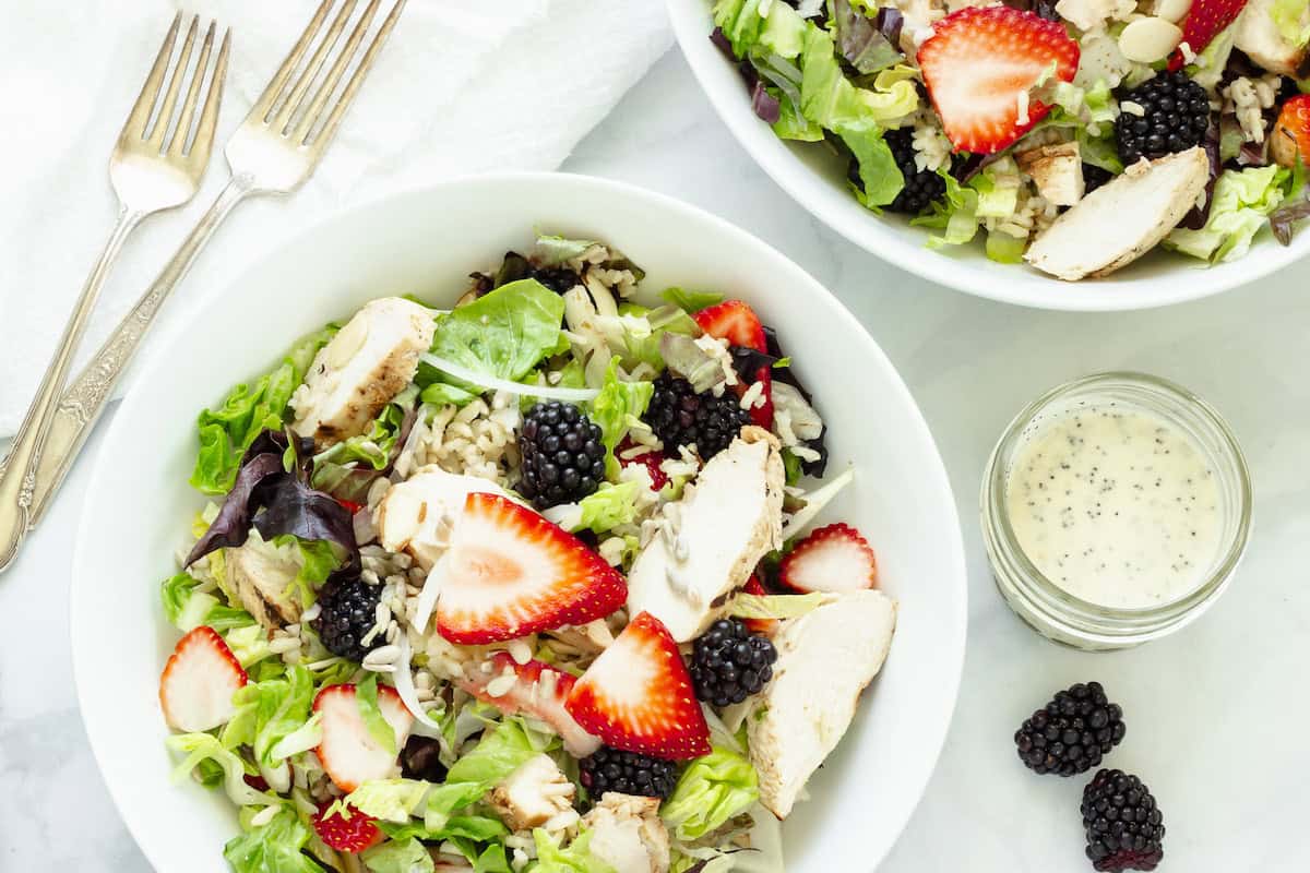 Two summer chicken and rice bowls with forks and a white napkin. Fresh blackberries and a small jar of salad dressing are next to the bowls.