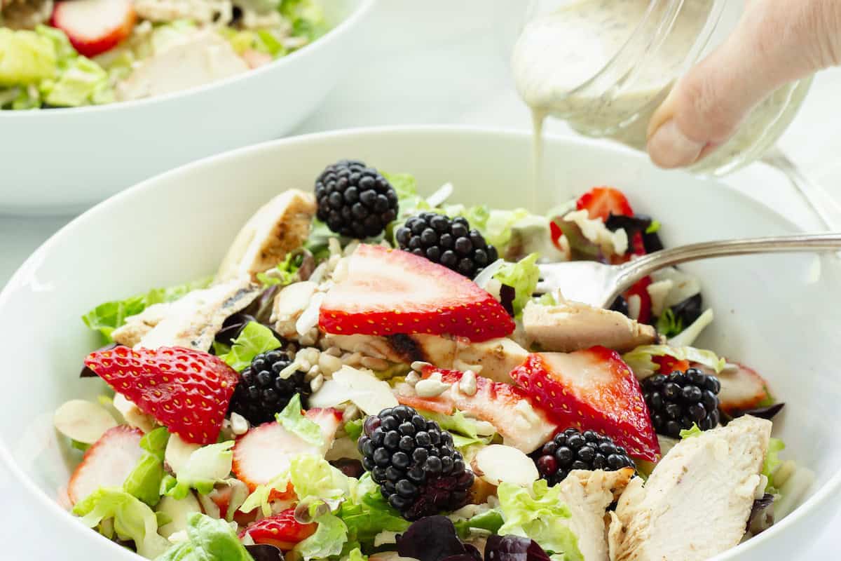 A chicken and rice bowl topped with fresh strawberries and blackberries. A small jar of salad dressing is being poured over the salad.