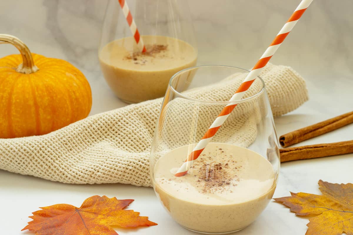 Two glasses of pumpkin pie smoothie with orange and white striped straws. Fall leaves and a mini pumpkin are in the background.