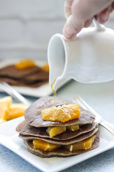 a plate of buckwheat pancakes with orange segments on top and a pitcher of syrup pouring over