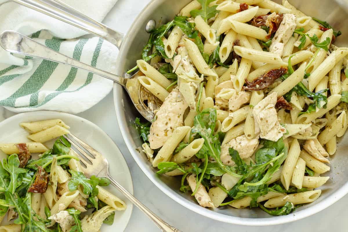 A skillet and a white plate of salmon pesto pasta on a white background. A green and white striped napkin is next to the pan.