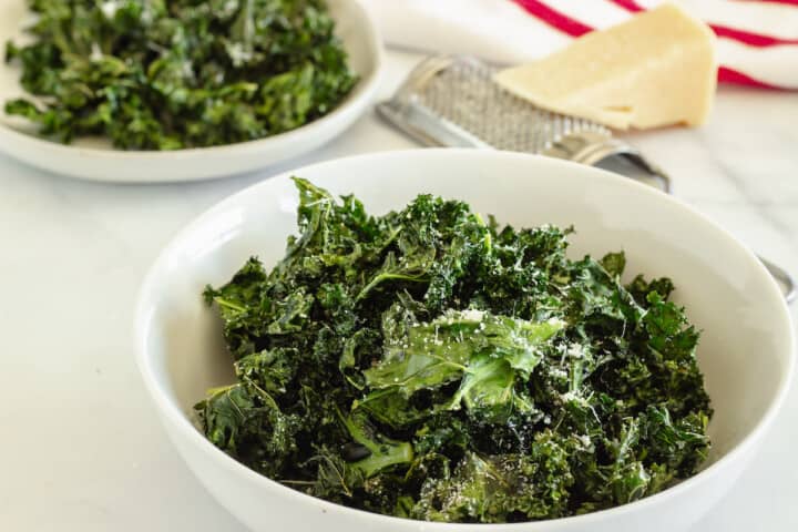 A white bowl full of air fryer kale chips. Parmesan cheese, a cheese grater, and a plate of kale chips are in the background.