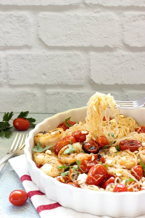 A casserole dish with garlic shrimp and tomatoes over shirataki noodles. A fork is lifting noodles out of the dish.