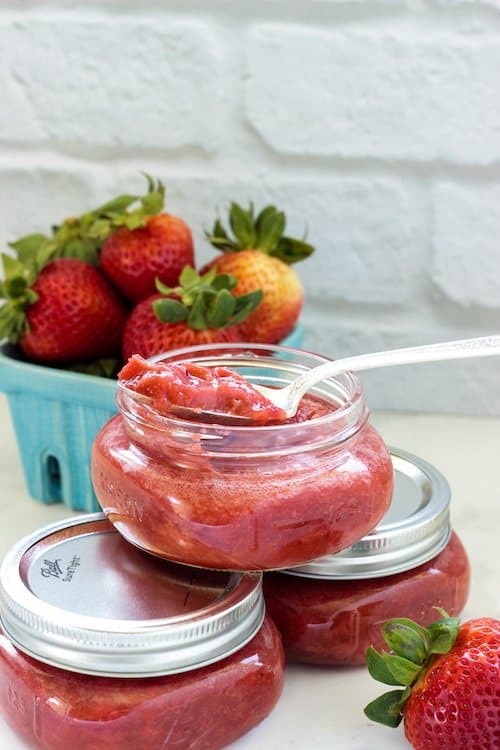 3 small jars of sugar-free strawberry rhubarb jam with a container of fresh strawberries in the background.