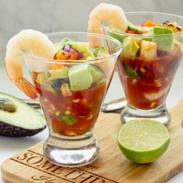 Two clear glasses with Mexican shrimp cocktail on a wooden board. A lime half and avocado half are next to the glasses.