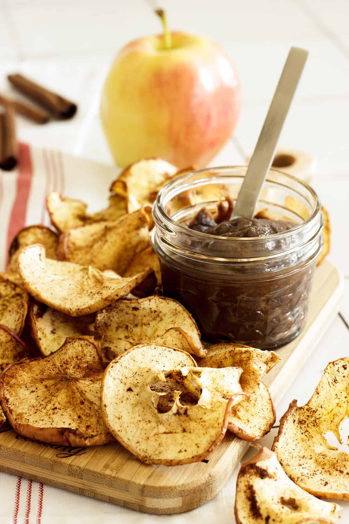 A pile of air fryer apple chips on a board with a small jar of chocolate sauce next to the chips.  An apple and cinnamon sticks in the background